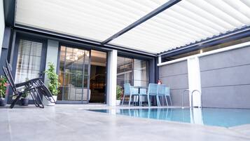 A modern outdoor pool area with a covered patio, seating, and a clear view into the building.