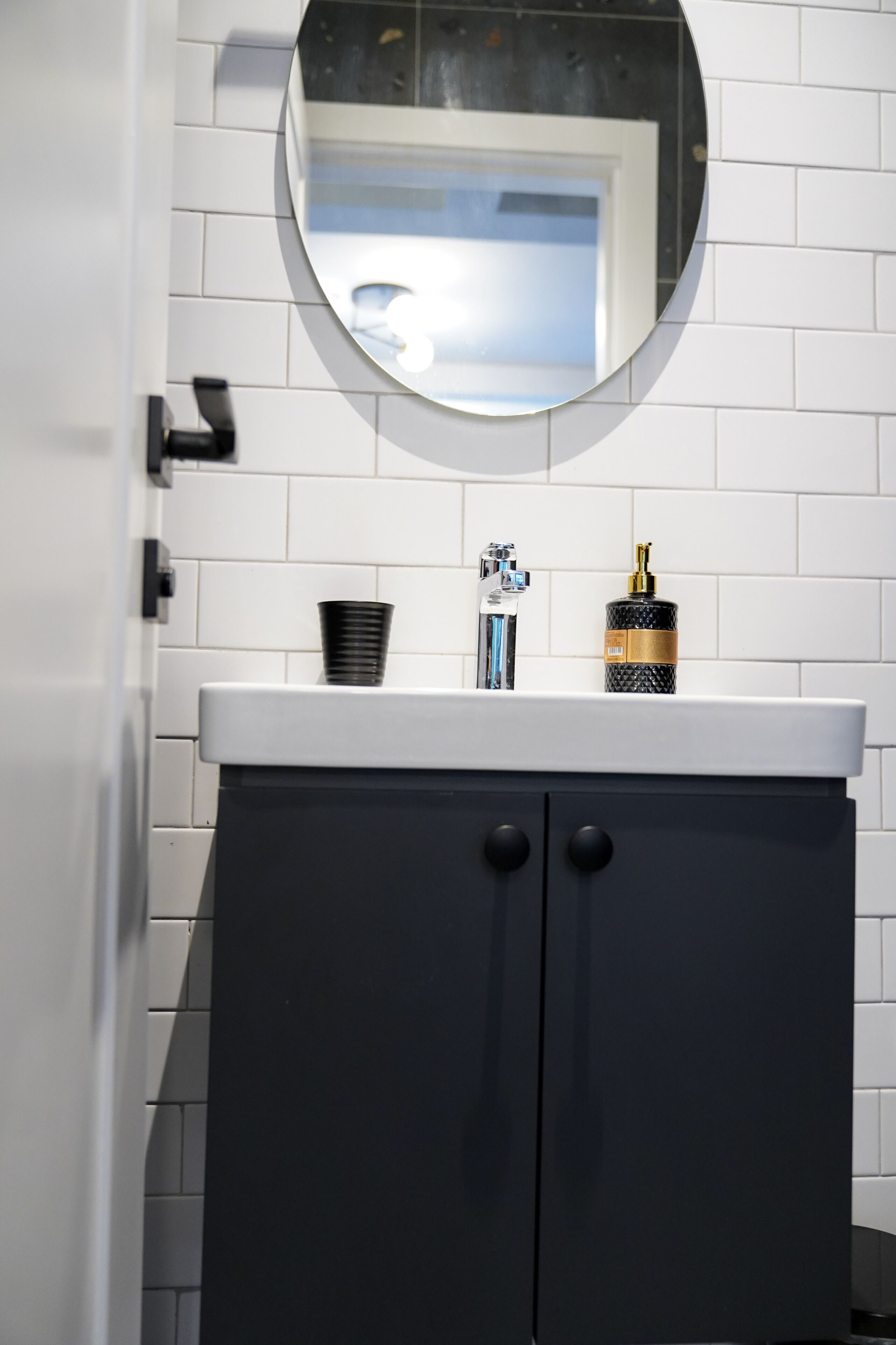 A bathroom with a white tiled wall, a round mirror, a black cabinet with two drawers, and a white countertop.