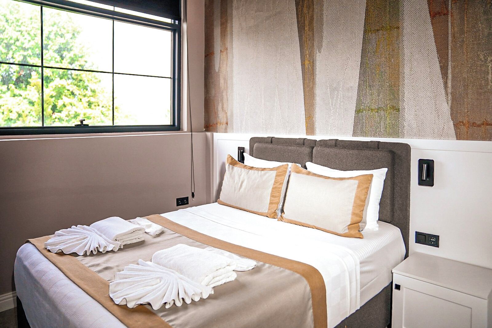 Un letto rifatto con lenzuola bianche e un tocco color beige, una finestra con vista sul verde e una parete rivestita con tessuto strutturato.