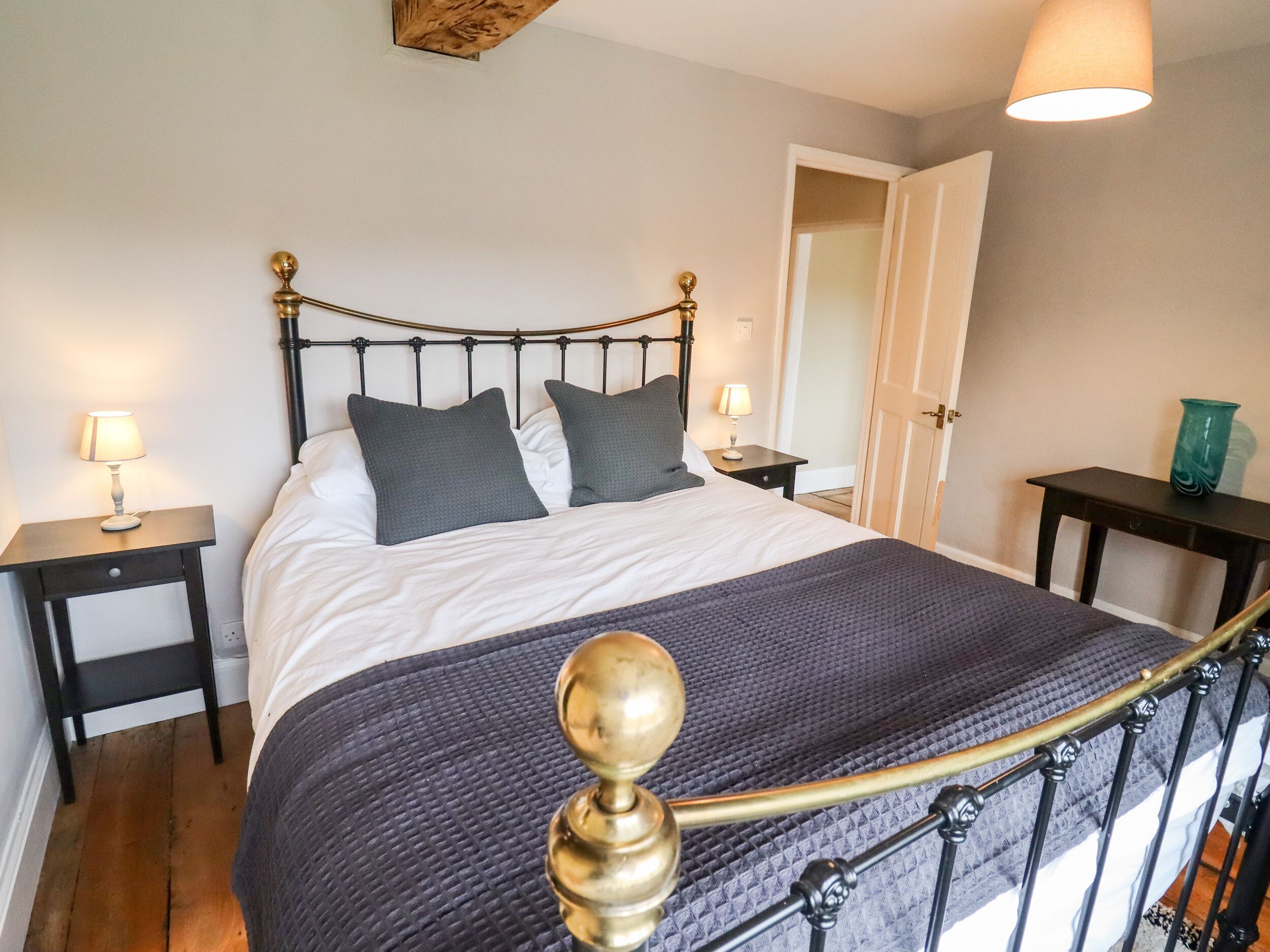 Una cama bien hecha con cabecera, dos mesitas de noche con lámparas, y una puerta que da a otra habitación.