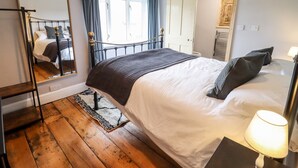 A bedroom with a bed, a bedside table with a lamp, a wooden floor, a window with curtains, a door, and a mirror on the wall. - Ley Mill Farm (Westbury-on-Severn)