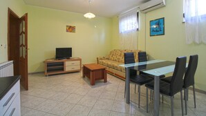 A living room with a sofa, a TV on a wooden stand, a glass dining table with chairs, and a tiled floor.