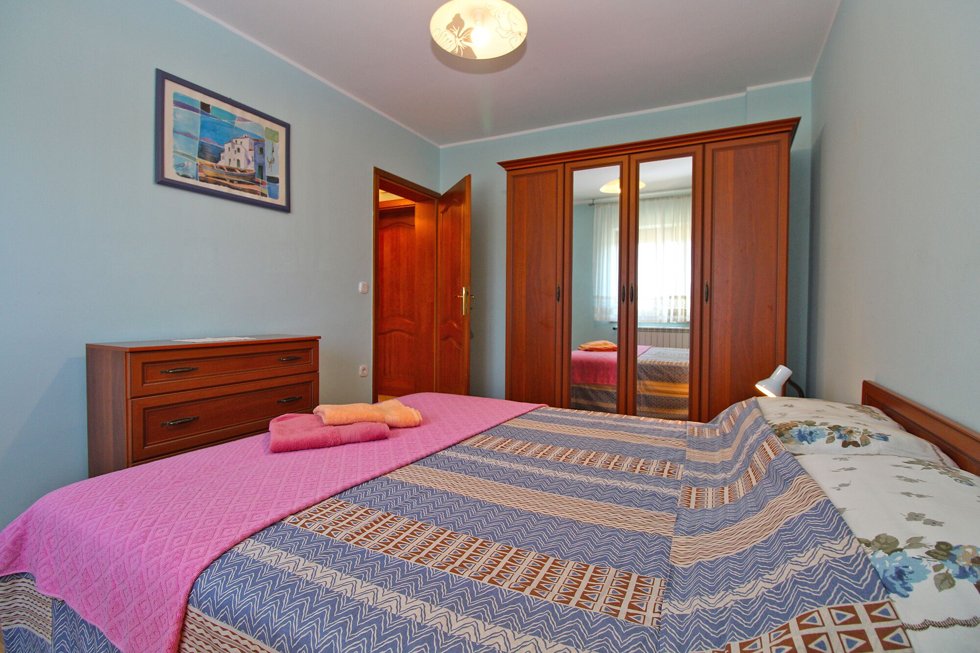 A bedroom with a bed, a wooden wardrobe, a dresser, a pink towel, and a framed picture on the wall.
