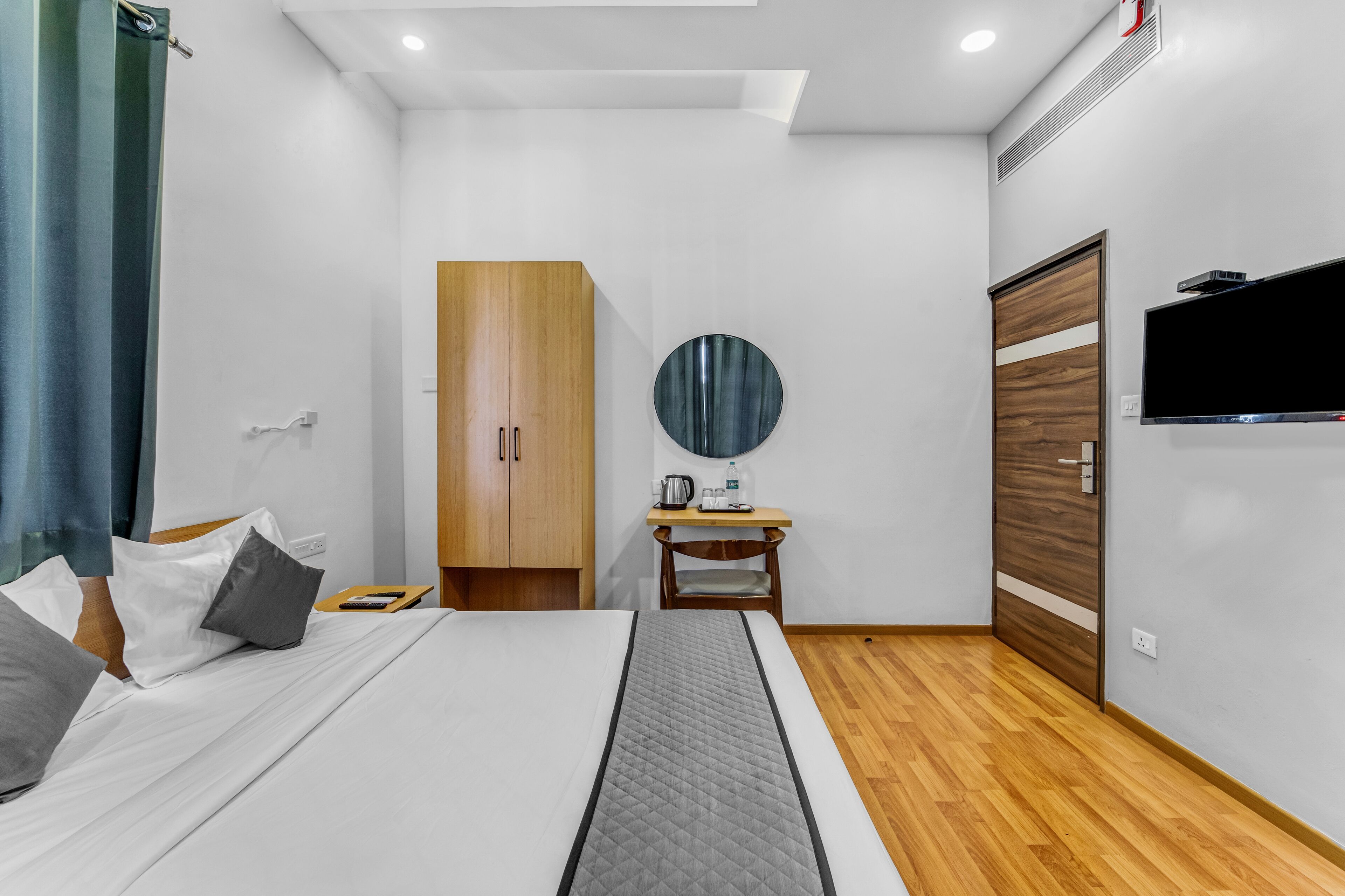 Ein modernes Hotelzimmer mit einem Bett, einem Holzschrank, einem kleinen Schreibtisch mit Stuhl, einem runden Spiegel und einem an der Wand befestigten Fernseher.