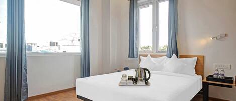 Una habitación de hotel con una cama grande, una mesita de noche con tetera y tazas, y una ventana con cortinas.