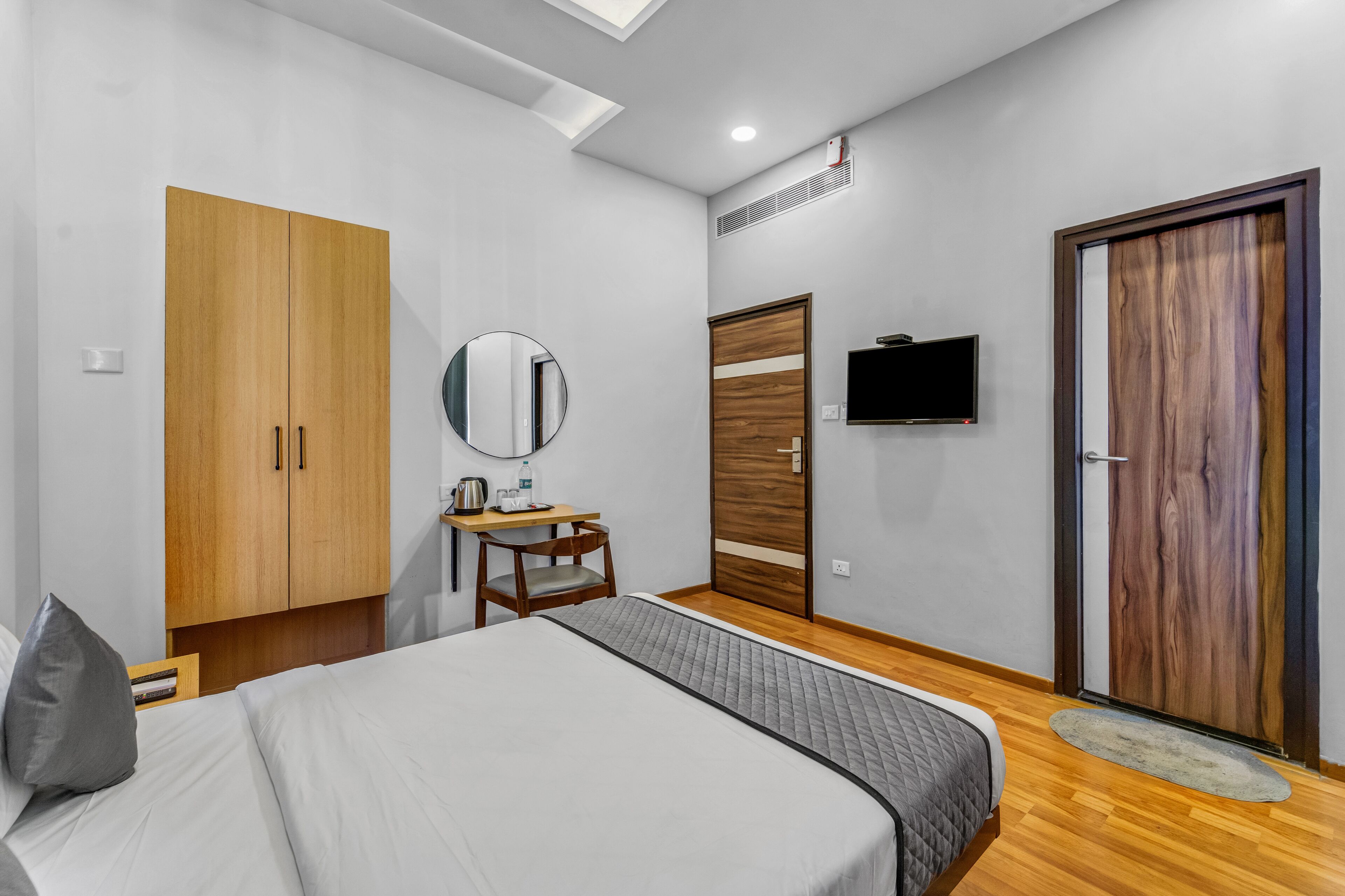 A modern bedroom with a bed, a wooden wardrobe, a small desk with a chair, a mirror, a television, and a door with a wooden panel.