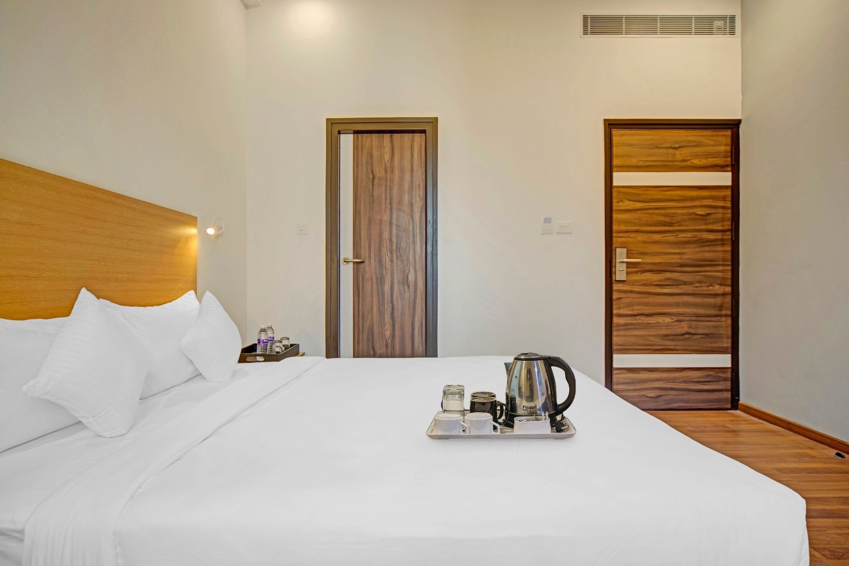 Habitación de hotel con una cama grande, cabecera de madera, dos puertas y una bandeja con tetera y tazas.