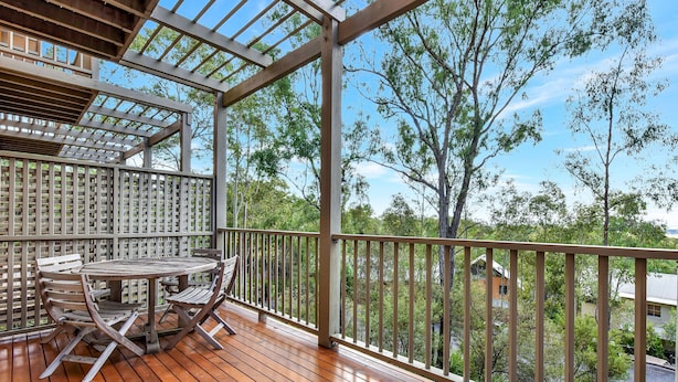 A wooden deck with a table and chairs, surrounded by a metal railing, overlooking a residential area with trees and houses.