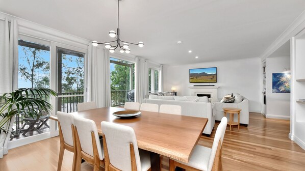 A modern living room with a wooden dining table, white sofas, and a large window with a view of trees.
