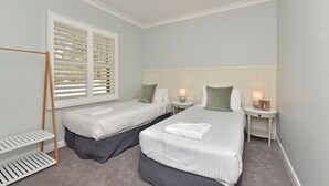 Habitación de hotel con dos camas individuales, una ventana con persianas, una mesita de noche con lámpara y un estante pequeño.