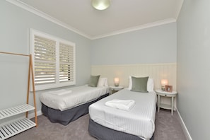 A hotel room with two single beds, a window with blinds, a bedside table with a lamp, and a small shelf.