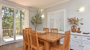 Un comedor con una mesa y sillas de madera, un jarrón con flores secas y una ventana con contraventanas blancas.