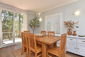 A dining area with a wooden table and chairs, a vase with dried flowers, and a window with white shutters.