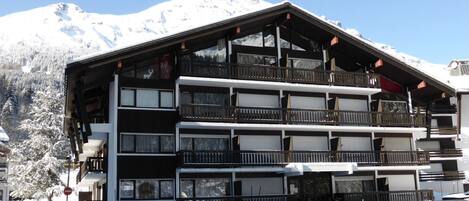 A multi-story hotel with balconies and large windows, set against a snowy mountain backdrop.