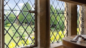 Une vue à travers une fenêtre ornée d’une grille à motifs losangés, dévoilant un jardin avec différentes plantes et une structure en pierre.