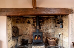 A stone fireplace with a black wood-burning stove, a wooden wheel, a black bucket, and a wicker basket filled with logs. - Little Gables (Broadway)
