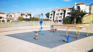 An outdoor exercise area with blue and orange exercise equipment, surrounded by a fence and residential buildings in the background. - Spacious 2-Room Apartment with Pool & Sports Facilities, Sleeps 4 – Le Barcarès (Le Barcarès)