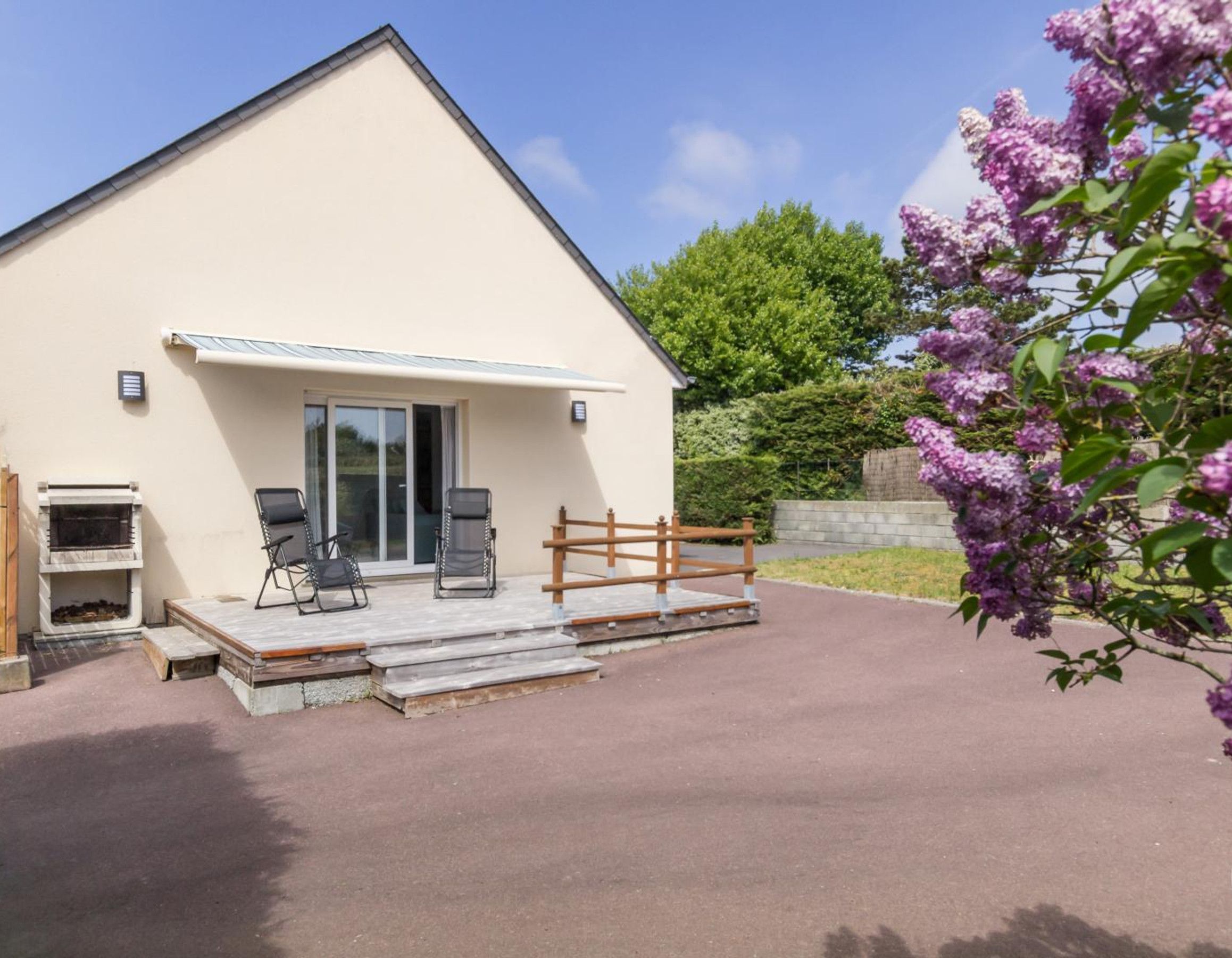 Ein modernes Gebäude mit flachem Dach, einer überdachten Terrasse mit zwei Stühlen, einem Grill und einem Holzzaun.
