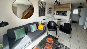 Un salon avec un canapé gris, un miroir rond, un coin repas avec une petite table et des chaises, et un coin cuisine avec un réfrigérateur et un luminaire suspendu.