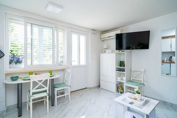 Una cocina luminosa y moderna con muebles blancos, una zona de comedor con una mesa redonda y sillas, un televisor de pantalla plana montado sobre los armarios, y una ventana con vistas a zonas verdes.