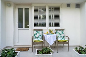 Un pequeño espacio al aire libre con sillas de mimbre y una mesa, una puerta blanca y una ventana con persianas.