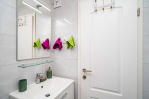 Un baño con un lavamanos blanco, un espejo y una puerta. Hay tres toallas colgadas en la pared.