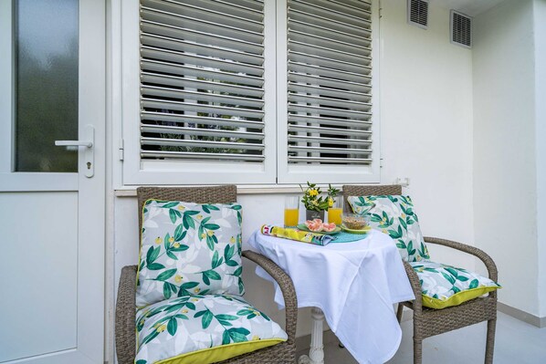 A wicker chair with a floral cushion, a small table with a white tablecloth, and a window with white shutters.