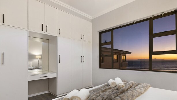 A bedroom with a large window, a bed with rolled towels, a mirror, and white cabinets. - Jacquelena Hof 9 (Cape Town)
