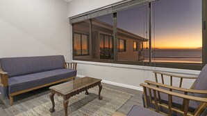 A room with a sofa, a wooden chair, a small table, and a large window with a view of a building and sunset. - Jacquelena Hof 9 (Cape Town)