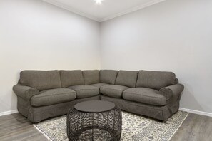 Uma sala de estar com canto, um sofá modular cinza, uma mesa de centro de vime redonda e um tapete estampado.