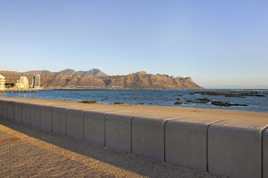 A coastal road with a concrete barrier, a beach, and mountains in the background.