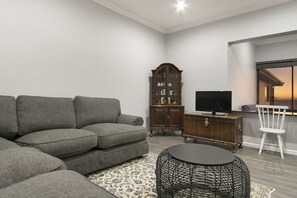 Una sala de estar con un sofá seccional gris, un mueble de madera, un televisor, una mesa de centro de mimbre y una silla blanca.