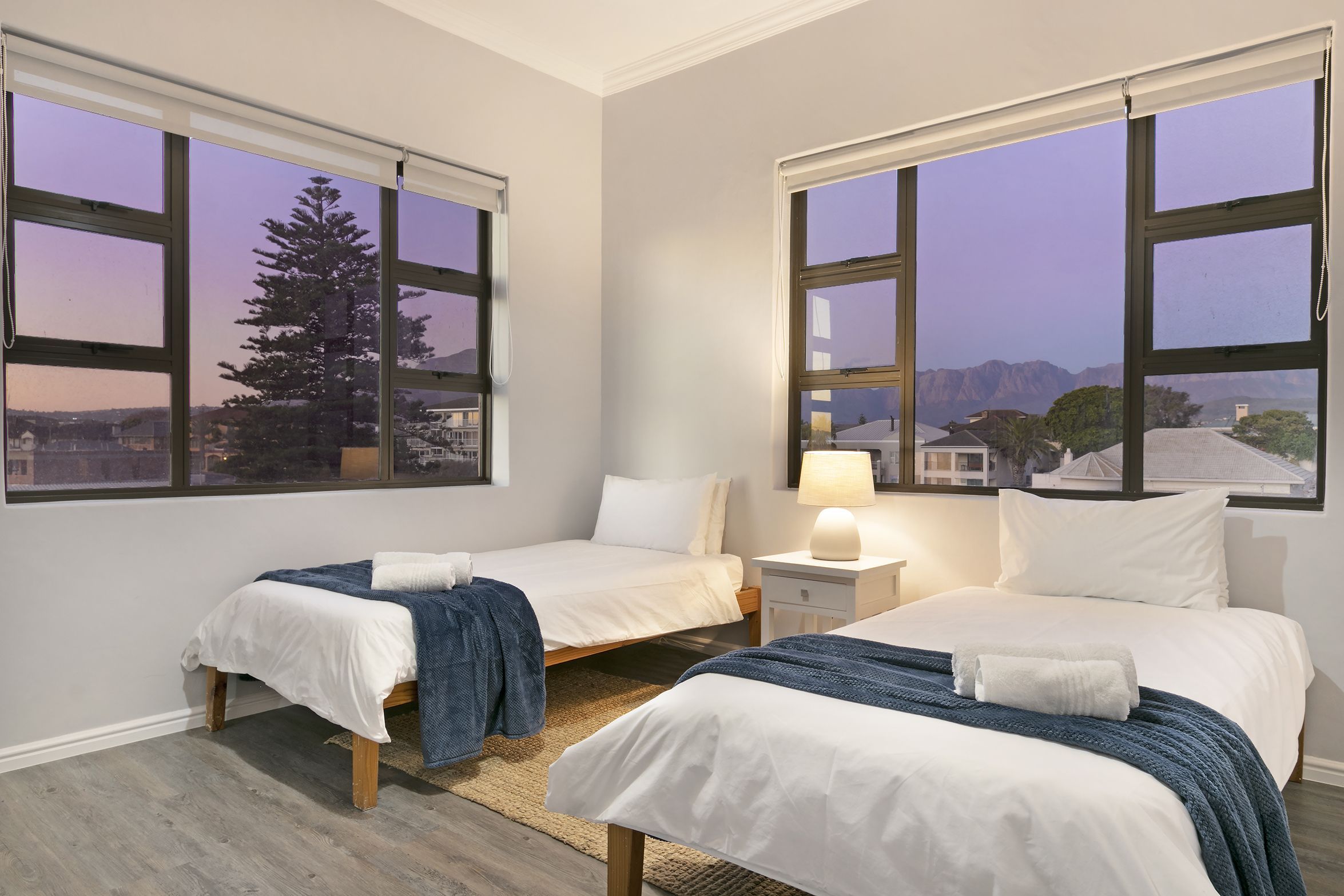 A bedroom with two beds, a nightstand with a lamp, and large windows with a view of trees and buildings.