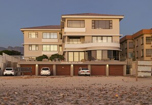 Um edifício residencial de vários andares com uma placa 'JACQUELENA HOF'. Três carros estão estacionados em frente e o edifício possui várias varandas.