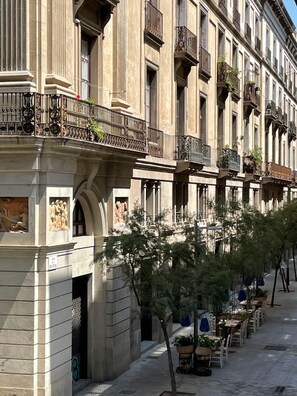 A row of traditional European-style buildings with balconies, a cobblestone street, and outdoor seating for cafes. - Som Nit Port Vell (Barcelona)