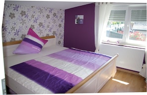 A bedroom with a bed, a purple wall, a window with blinds, and a radiator.