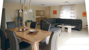 A modern living room with a dining area, featuring a wooden dining table, leather sofas, and a large window with curtains. - Vacation home for max. 6 persons in Duhnen (Cuxhaven)