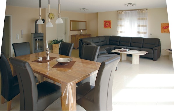A modern living room with a dining area, featuring a wooden dining table, leather sofas, and a large window with curtains.