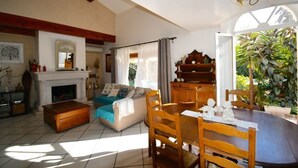 A cozy living room with a fireplace, a sofa, a dining table with chairs, and a large window with plants.