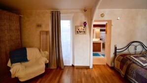 A cozy bedroom with a wooden floor, a bed with a patterned blanket, a chair with a blanket, and a doorway leading to a bathroom.