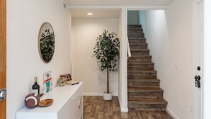 Un pasillo con una escalera de madera, una planta en maceta, un mostrador blanco con espejo y un cuadro enmarcado.