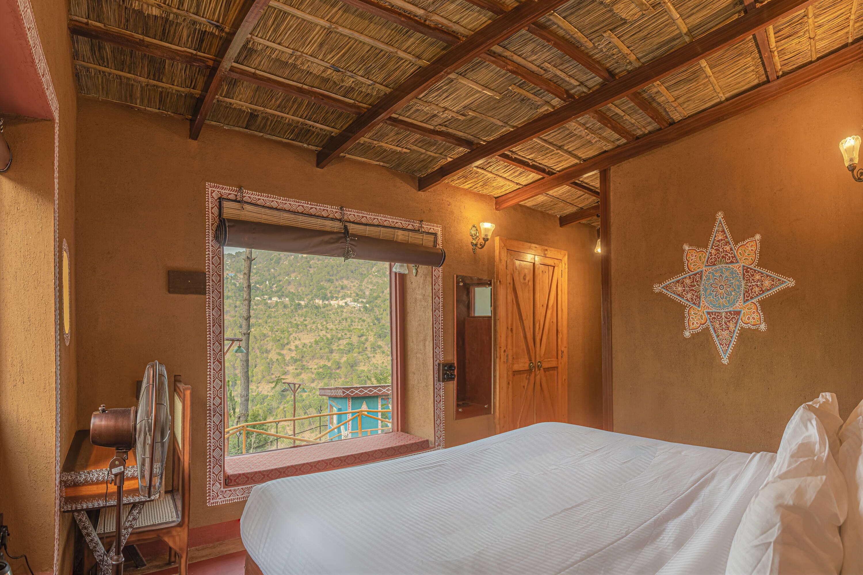 A room with a bed, a window with a view of greenery, a wooden door, and a decorative wall piece.