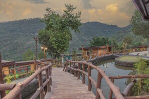 A wooden bridge leading to a building with traditional architecture, surrounded by lush greenery and a mountainous backdrop. - A Kasauli Ggaon by Echor (Kasauli)