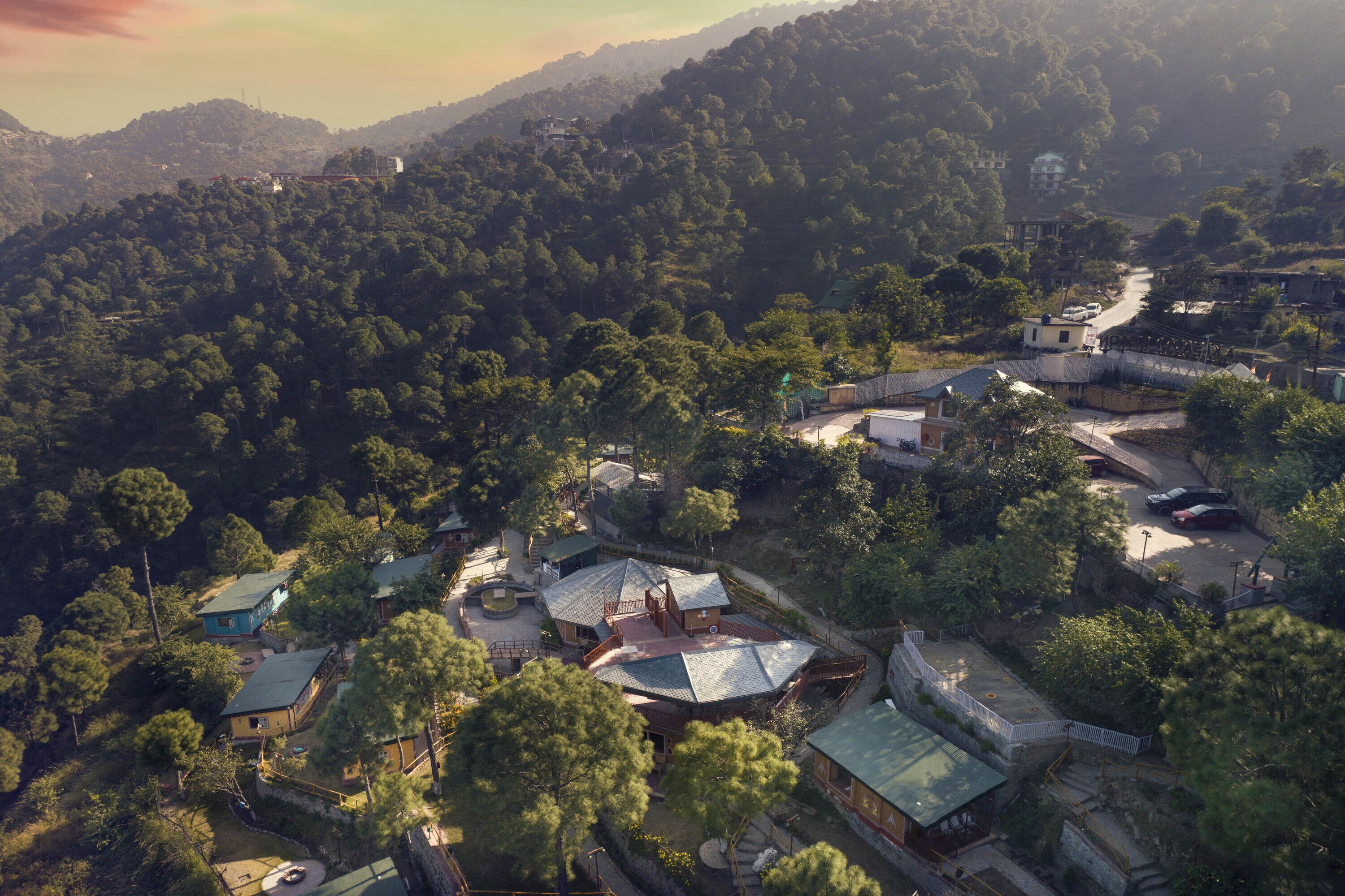 A residential area with houses surrounded by dense forest, a winding road, and a distant hill.