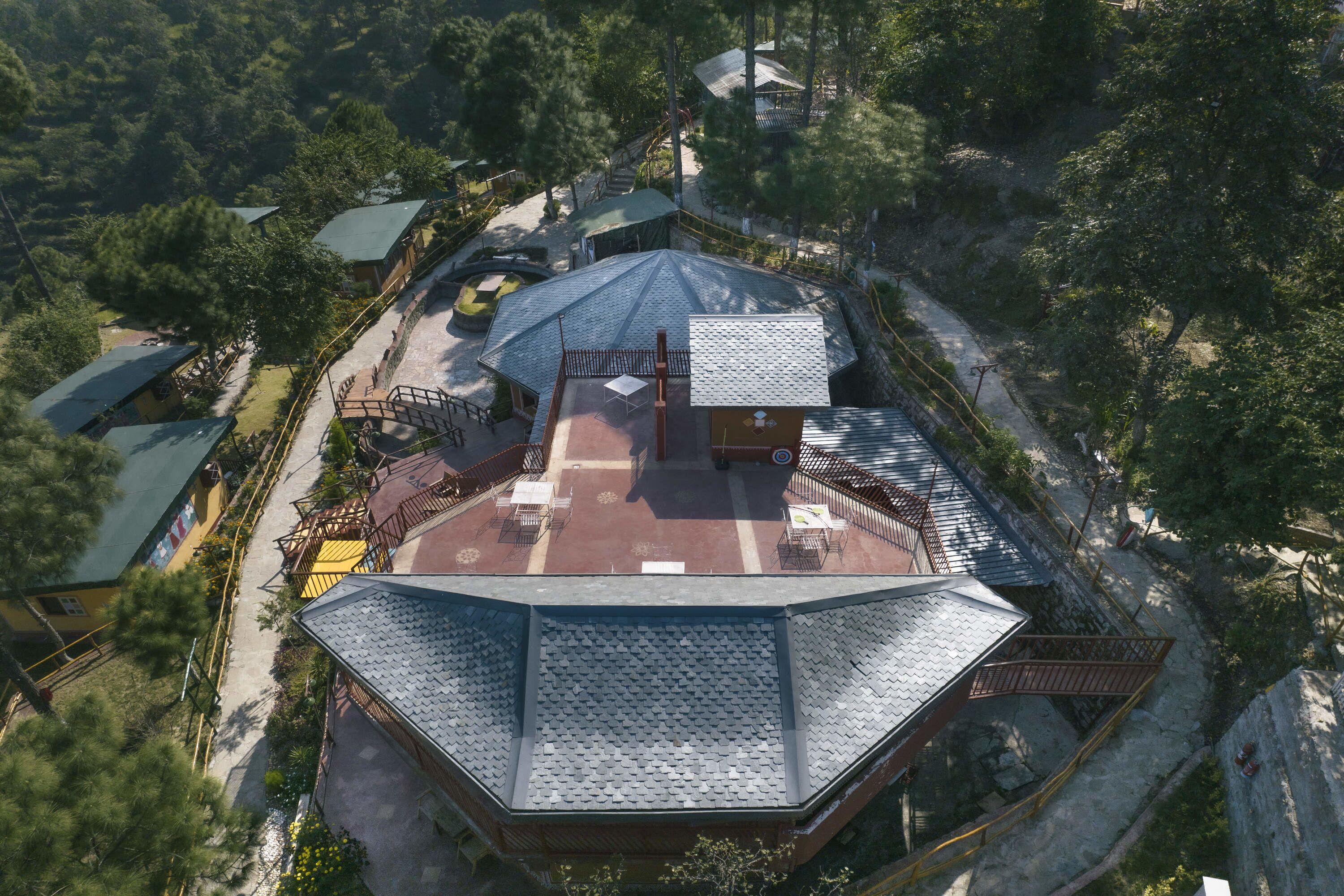 An aerial view of a building with a sloped roof, surrounded by trees and a small courtyard with a table and chairs.