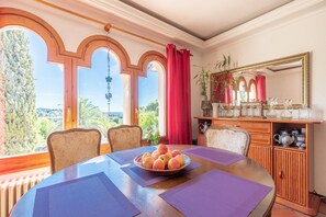 A dining area with a round table, chairs, a bowl of fruit, a cabinet with glassware, and a large window with a view.
