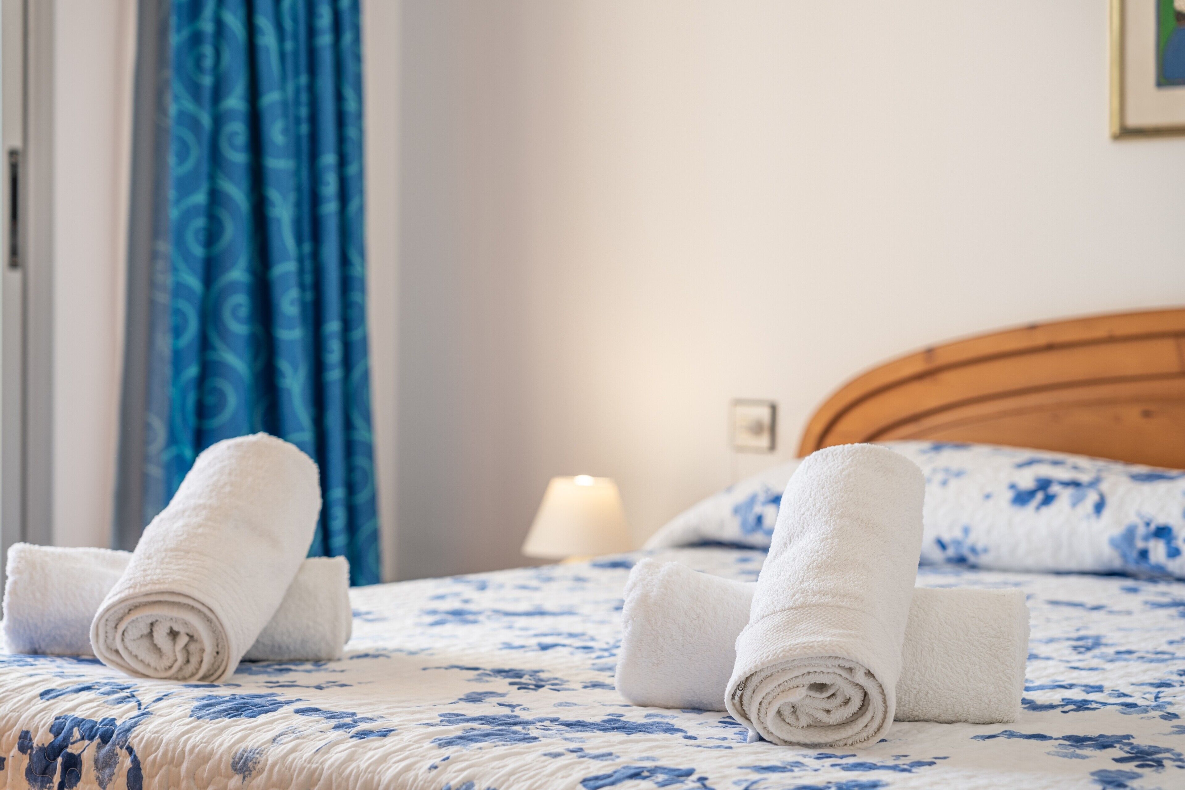 Una cama bien tendida con toallas blancas enrolladas, un cabecero de madera y una colcha azul estampada.