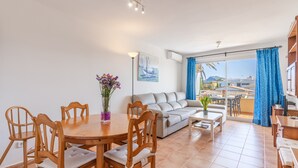 A living room with a dining table, chairs, a sofa, and a balcony with blue curtains. - Holiday Apartment "Veleta ll" close to Beach with Sea View, Shared Pool & Wi-Fi (Pollença)