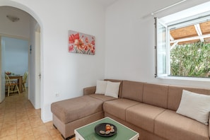 A living room with a beige sofa, a green coffee table, and a window with a view of greenery. - Apartment "Margaret House" with Wi-Fi, A/C, Terrace & Garden (Paros)