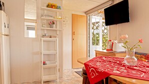 A kitchen with a white refrigerator, a wooden table with a red tablecloth, a TV mounted on the wall, and a door leading to a balcony.